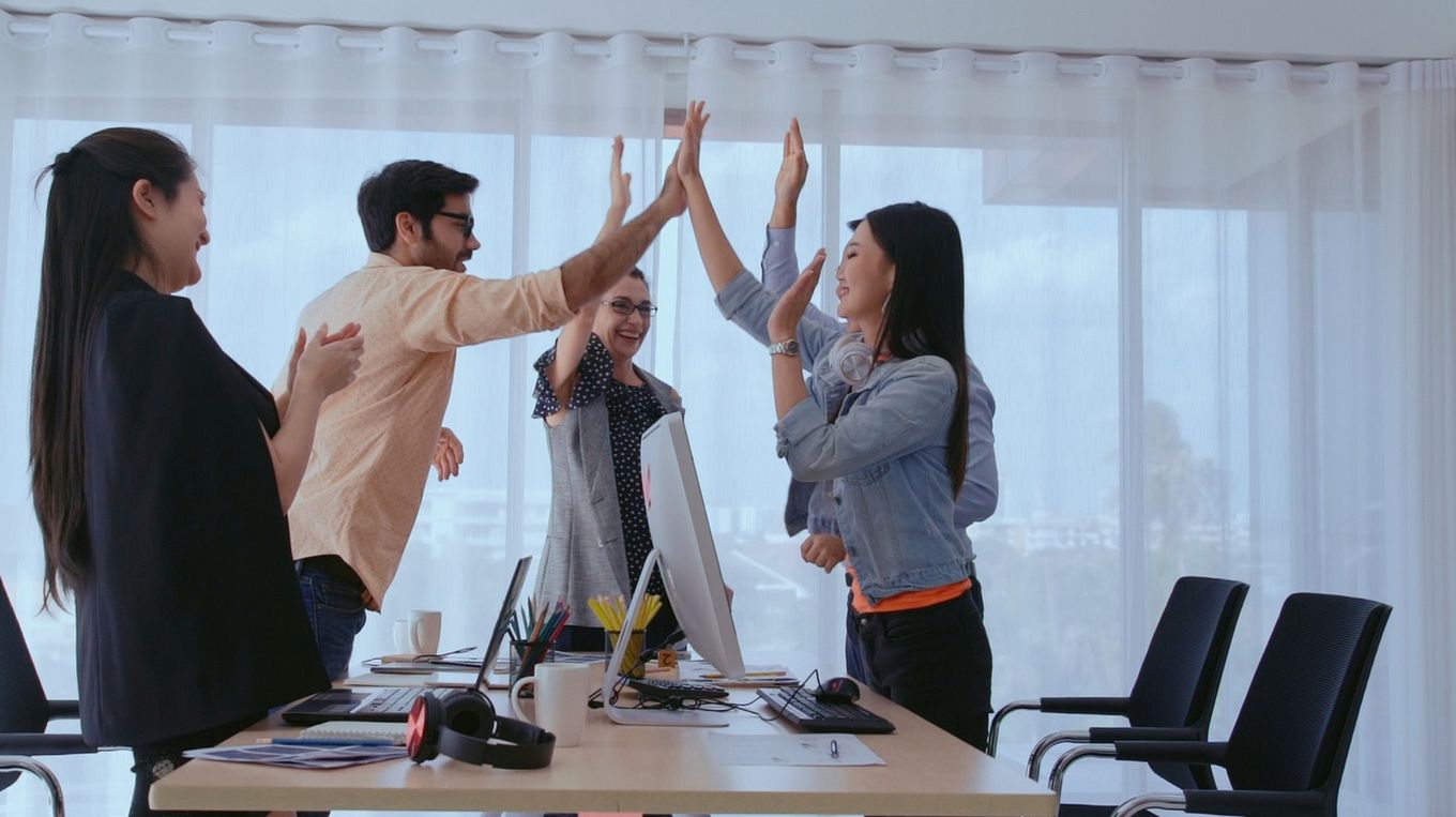 Na sliki skupina sodelavcev v pisarni proslavlja uspeh z dvigom rok in ploskanjem.